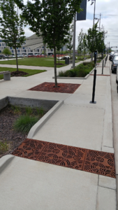 Cast iron trench drain grates in decorative Oblio pattern cuts across sidewalk to bioswale