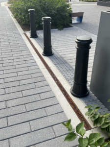 Cast iron trench drain grates with decorative Rain pattern alongside bollards on one side and grey pavers on the other