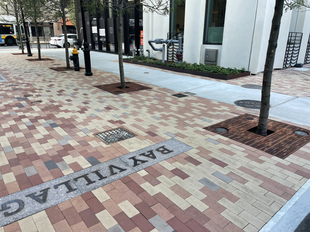 Cast iron tree grate with light hole cut-outs in linear Rain pattern, set in plaza of multicolored pavers