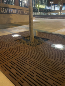 Cast iron tree grate with light hole cut-outs in linear Rain pattern, set in plaza of multicolored pavers