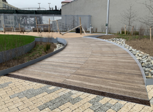 Cast iron trench drain grates with decorative Rain pattern, installed between wooden boardwalk on one side and blond/grey pavers on the other side.