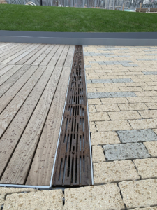 Cast iron trench drain grates with decorative Rain pattern, installed between wooden boardwalk on one side and blond/grey pavers on the other side.