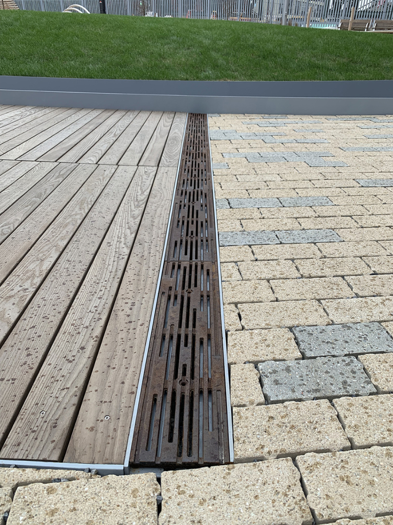 Cast iron trench drain grates with decorative Rain pattern, installed between wooden boardwalk on one side and blond/grey pavers on the other side.