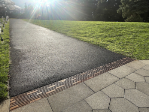 Cast iron trench drain grates with decorative Rain pattern, installed between asphalt walkway and grass on one side and hexagonal pavers on the other side.