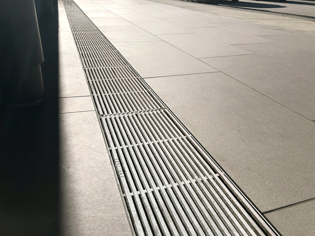 Cast aluminum trench drain grates in sleek, linear Que pattern, installed in concrete plaza.