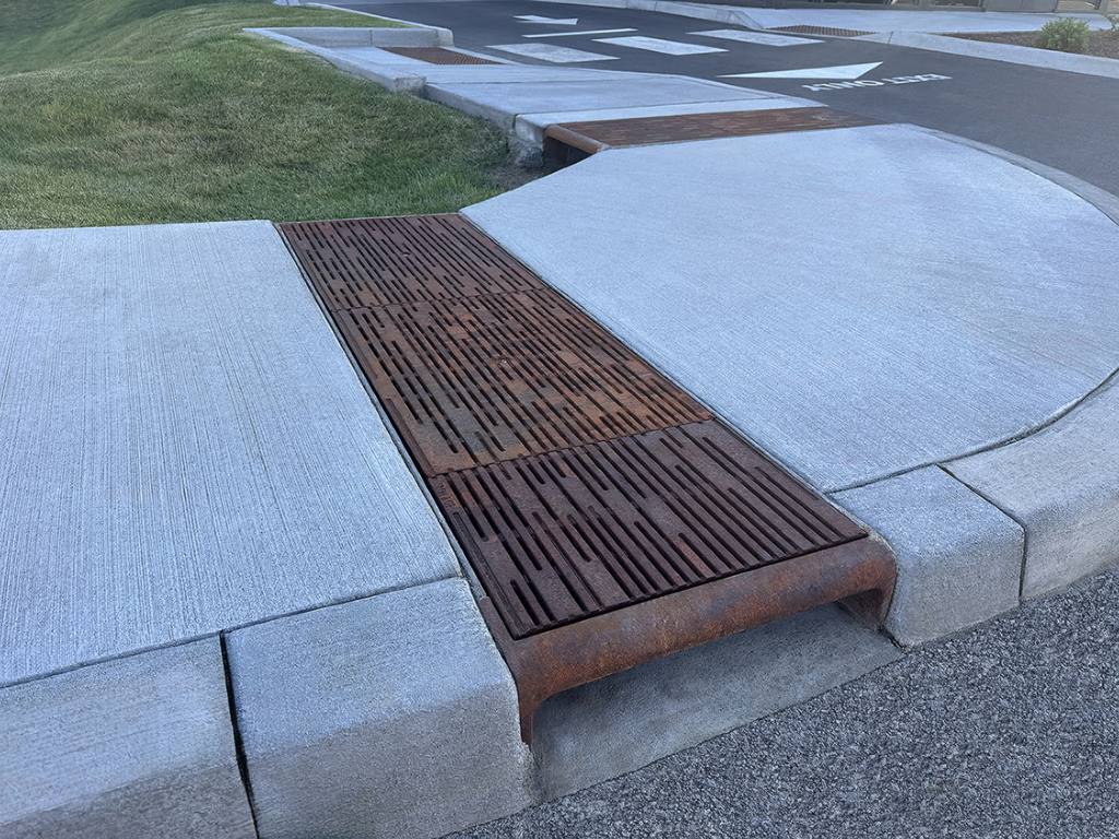 Cast iron curb inlet with decorative cast iron trench grates in Rain pattern by Iron Age Designs covering open runnel in sidewalk.