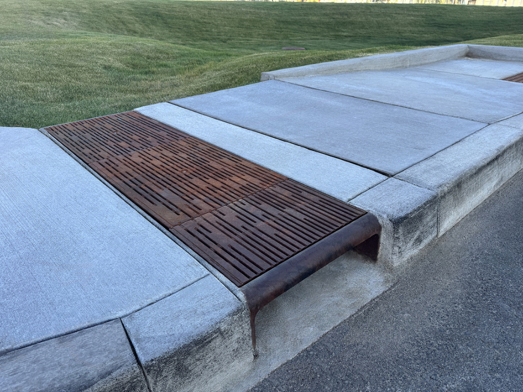 Cast iron curb inlet with decorative cast iron trench grates in Rain pattern by Iron Age Designs covering open runnel in sidewalk.