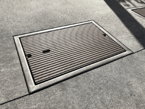 Cast iron utility cover with sleek, linear Que pattern. Installed with concrete surround.