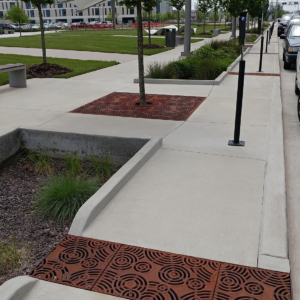 Cast iron trench drain grates in decorative Oblio pattern cuts across sidewalk to bioswale
