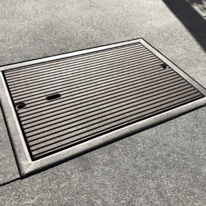 Cast iron utility cover with sleek, linear Que pattern. Installed with concrete surround.