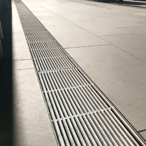 Cast aluminum trench drain grates in sleek, linear Que pattern, installed in concrete plaza.