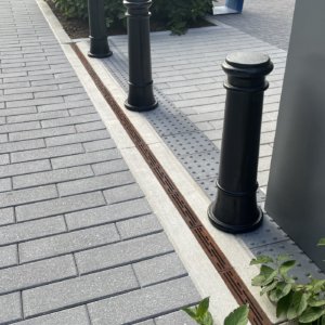 Cast iron trench drain grates with decorative Rain pattern alongside bollards on one side and grey pavers on the other