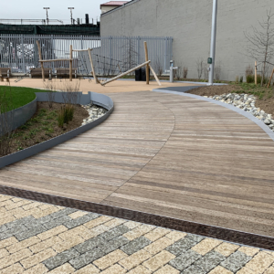 Cast iron trench drain grates with decorative Rain pattern, installed between wooden boardwalk on one side and blond/grey pavers on the other side.