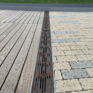 Cast iron trench drain grates with decorative Rain pattern, installed between wooden boardwalk on one side and blond/grey pavers on the other side.