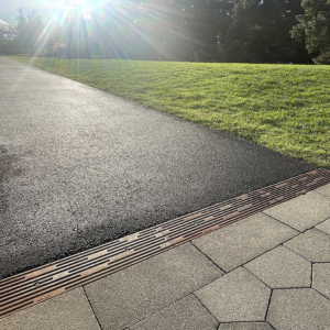 Cast iron trench drain grates with decorative Rain pattern, installed between asphalt walkway and grass on one side and hexagonal pavers on the other side.