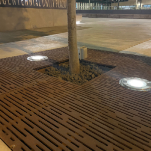 Cast iron tree grate with light hole cut-outs in linear Rain pattern, set in plaza of multicolored pavers