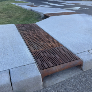 Cast iron curb inlet with decorative cast iron trench grates in Rain pattern by Iron Age Designs covering open runnel in sidewalk.